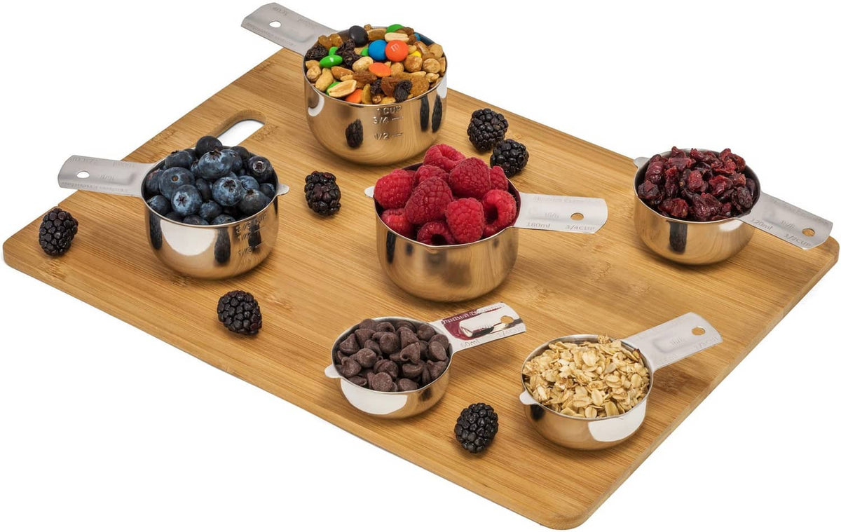 Stainless steel measuring cups on a board with berries and dry grains