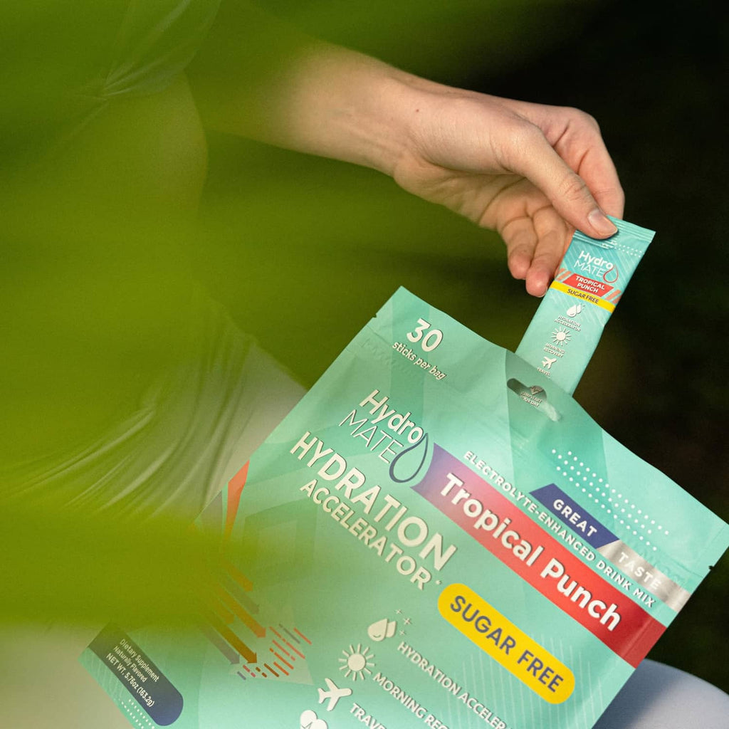 Hand holding HydroMate sugar-free Tropical Punch hydration packet, plus green pouch