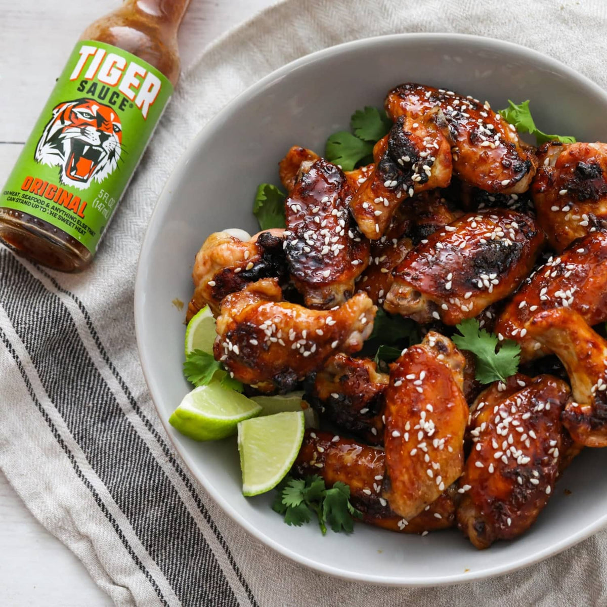 Hot sauce bottle beside plate of spicy wings with lime wedges and cilantro