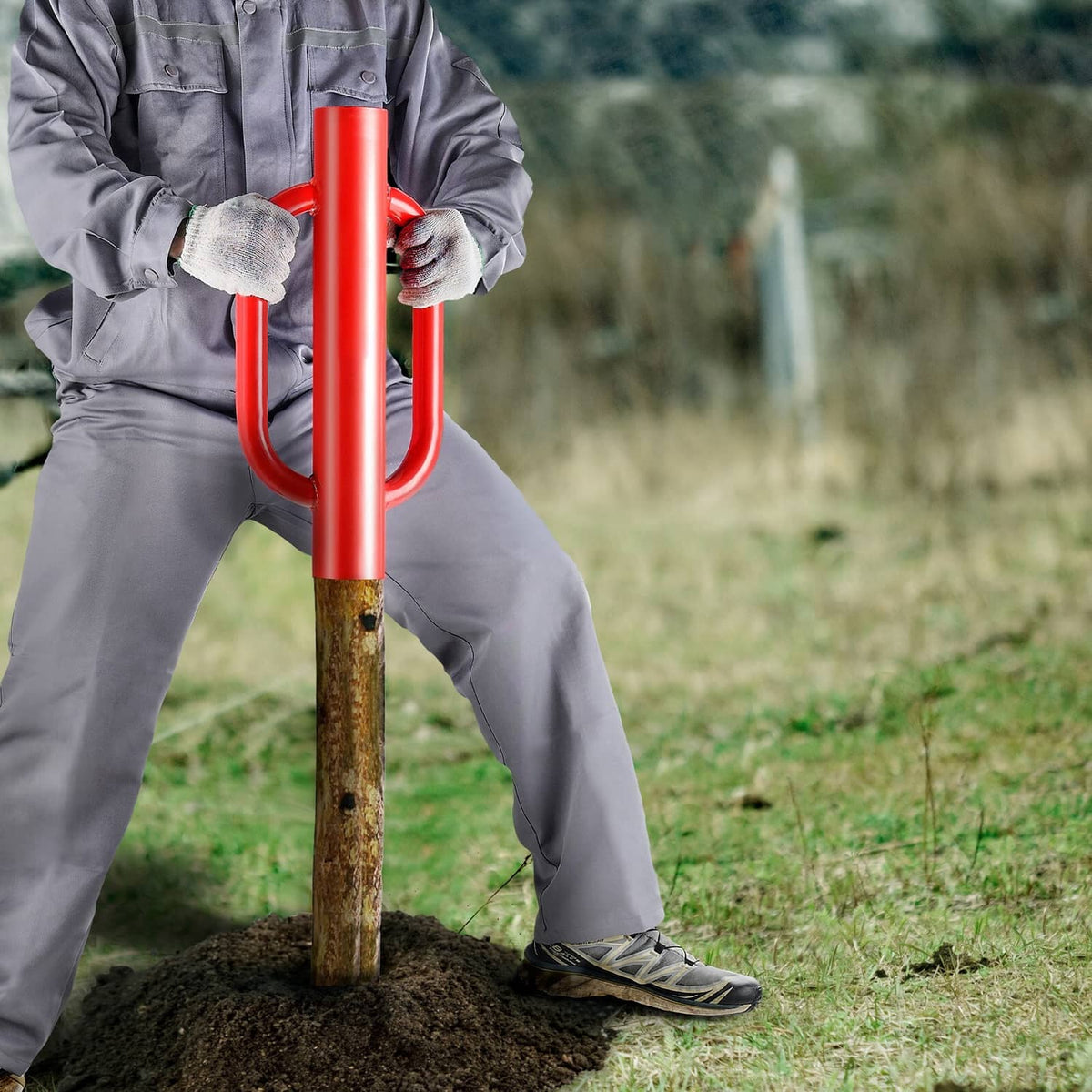 Red-handled fence post driver with U grips pounding a T-post into soil