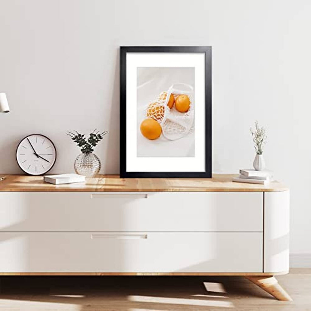 Black picture frame with mat, holding a photo of oranges in a net bag.