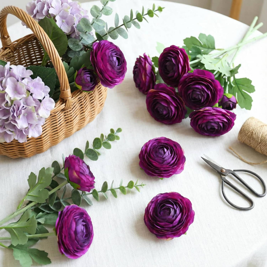 Purple artificial ranunculus flower heads with green leaves laid on a tabletop