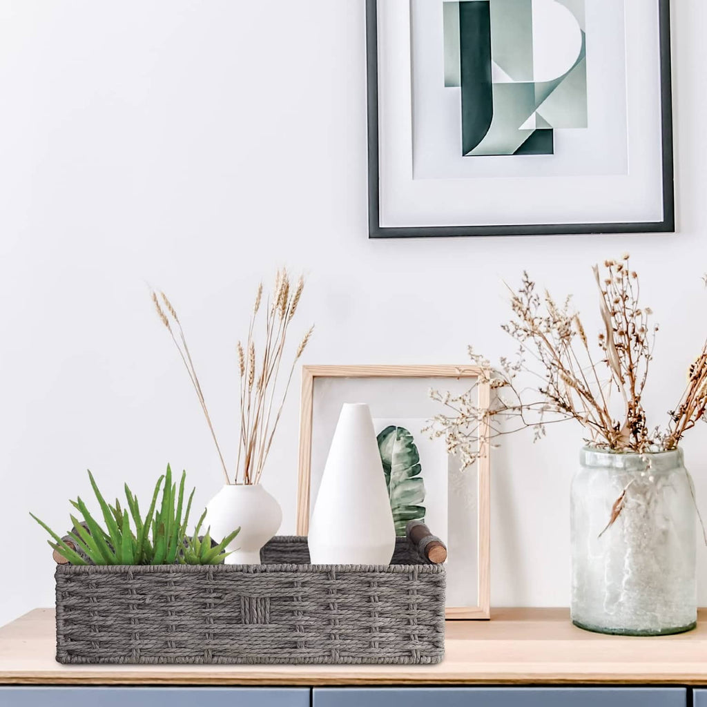 Grey woven storage basket on table with dried stems and green plant beside wooden handles.