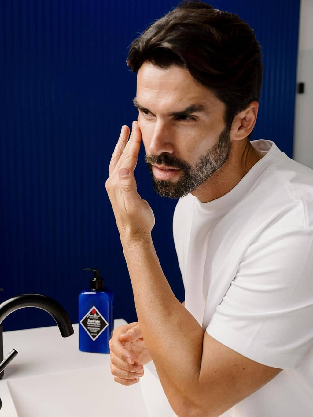 Man applying beard conditioner gel near jaw while holding blue bottle with pump