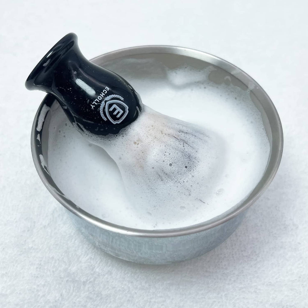 Men's black shaving brush head in metal bowl, visible lather and foam