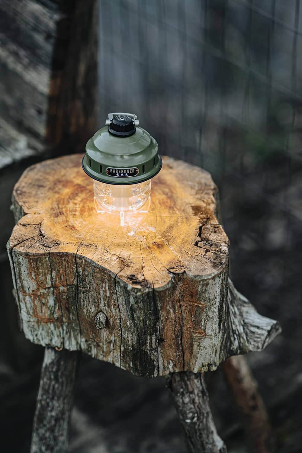 Olive drab camping lantern with warm glowing LED, resting on a cut wood log