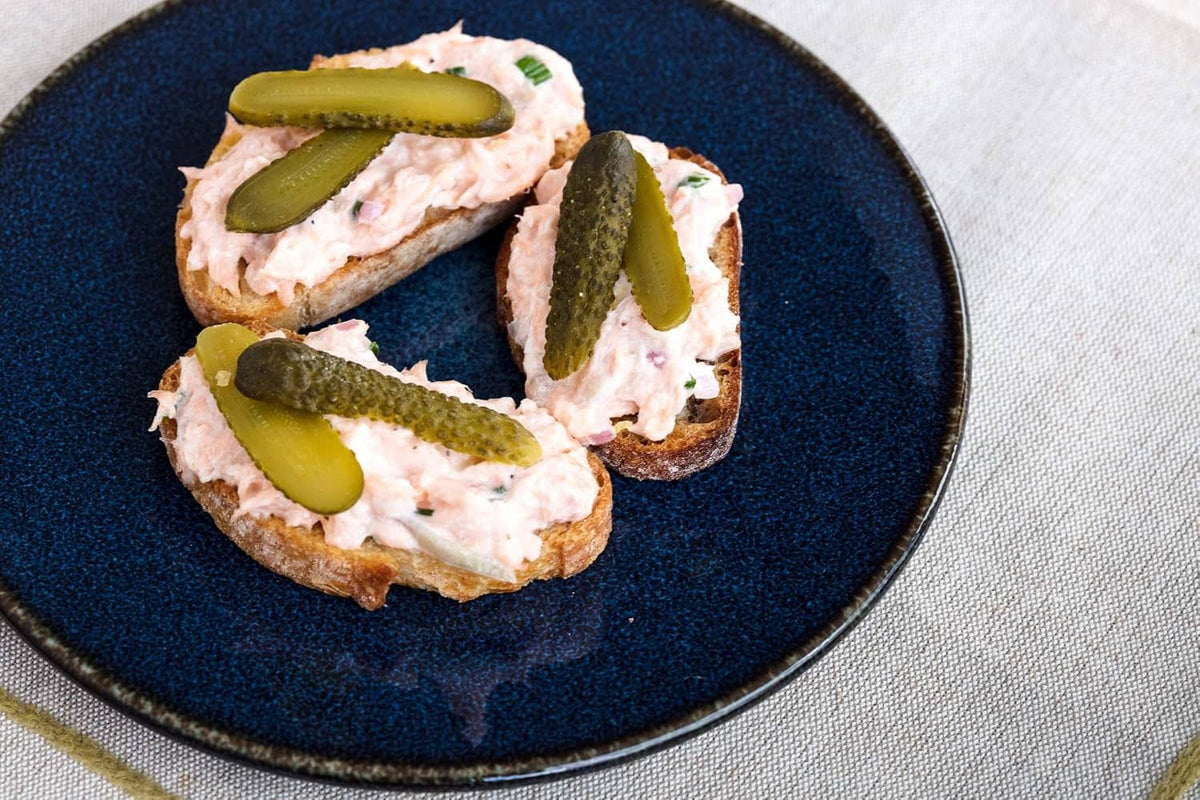 Open-faced bread bites topped with sliced green cornichons on a blue plate