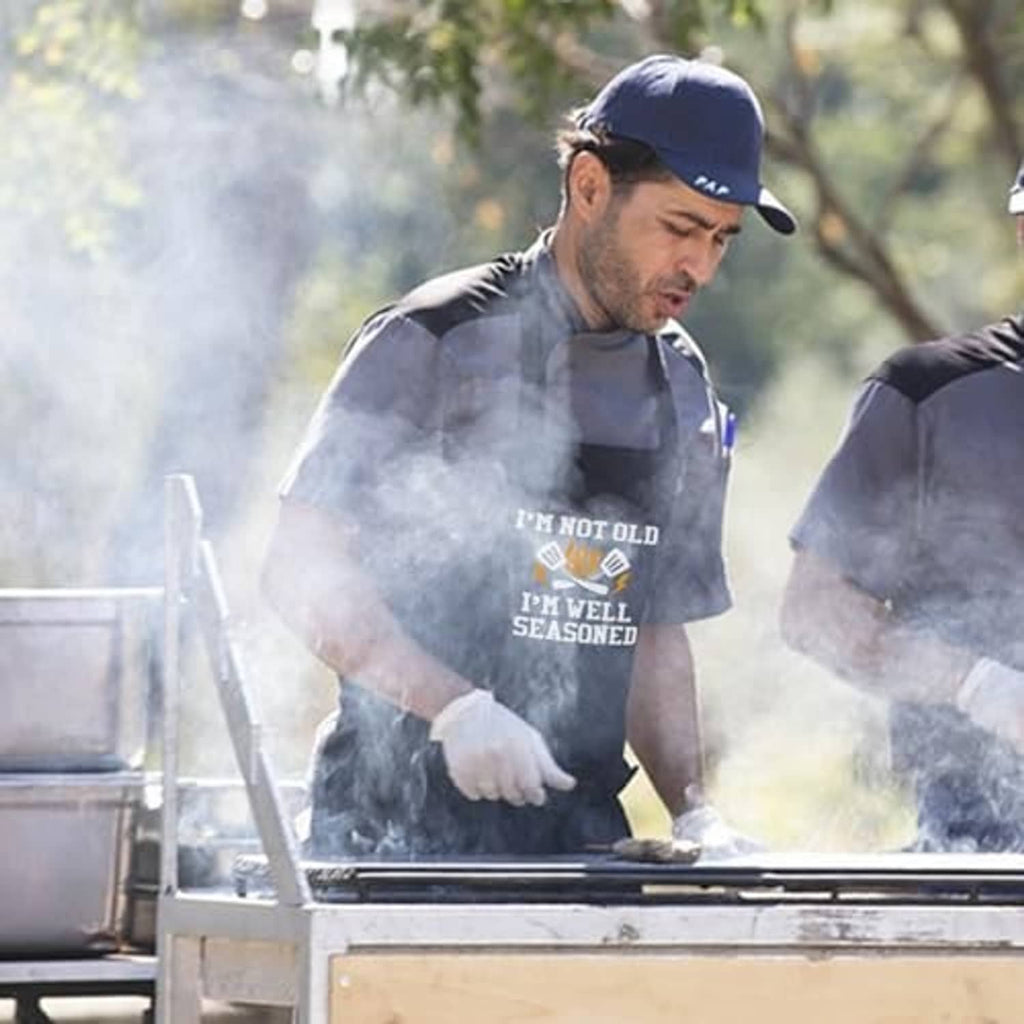 Men's black grilling apron with 'I'm well seasoned' print worn at an outdoor barbecue