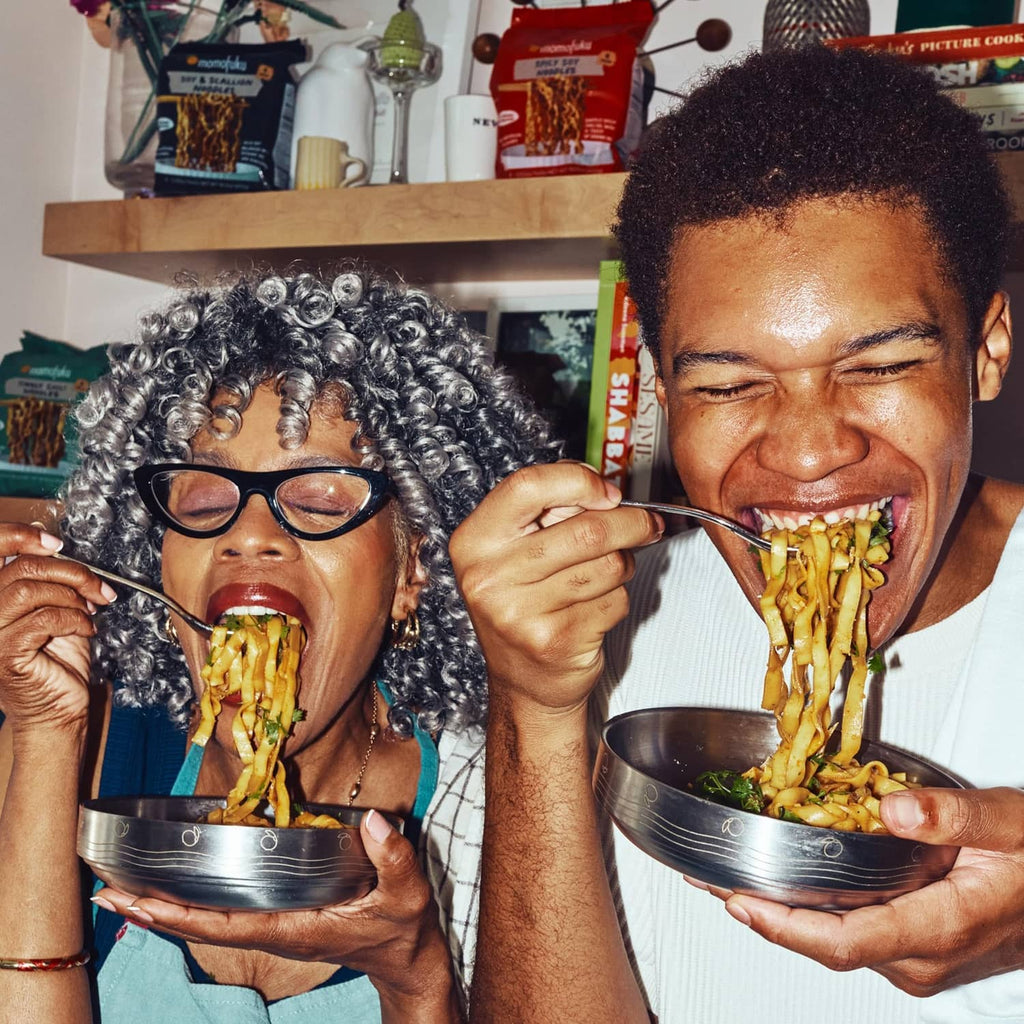 Two people eating saucy ramen noodles with serving bowls and product packs nearby