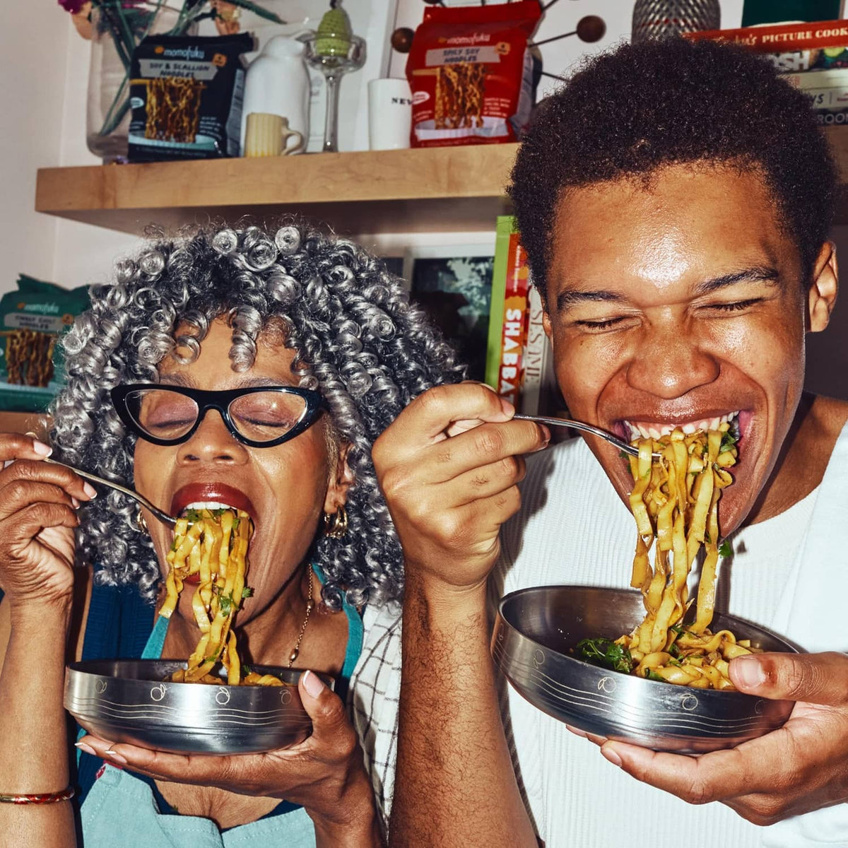 Two people eating saucy ramen noodles with serving bowls and product packs nearby