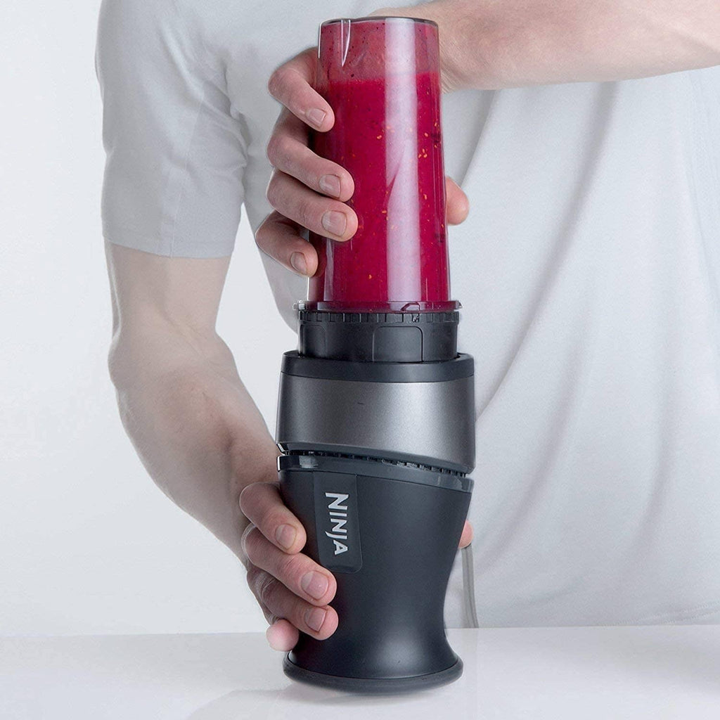 Clear blender cup with black lid, filled smoothie, held in hand for blending.