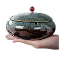 Turquoise ceramic ashtray with hinged lid and red knob, held in a hand