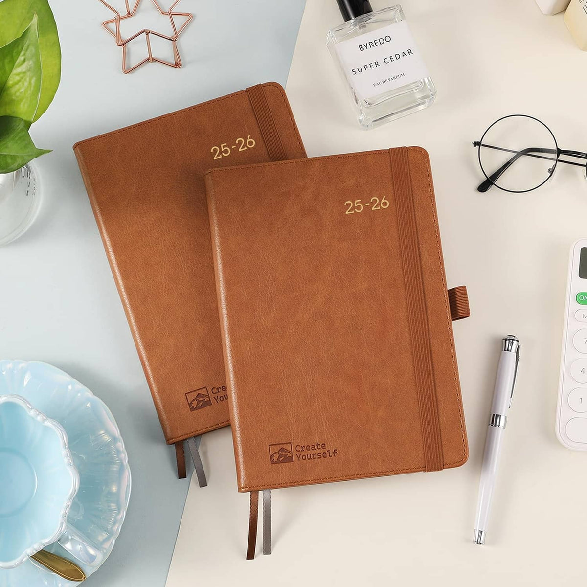 Two brown 25-26 planners with elastic bands, front covers angled on desk