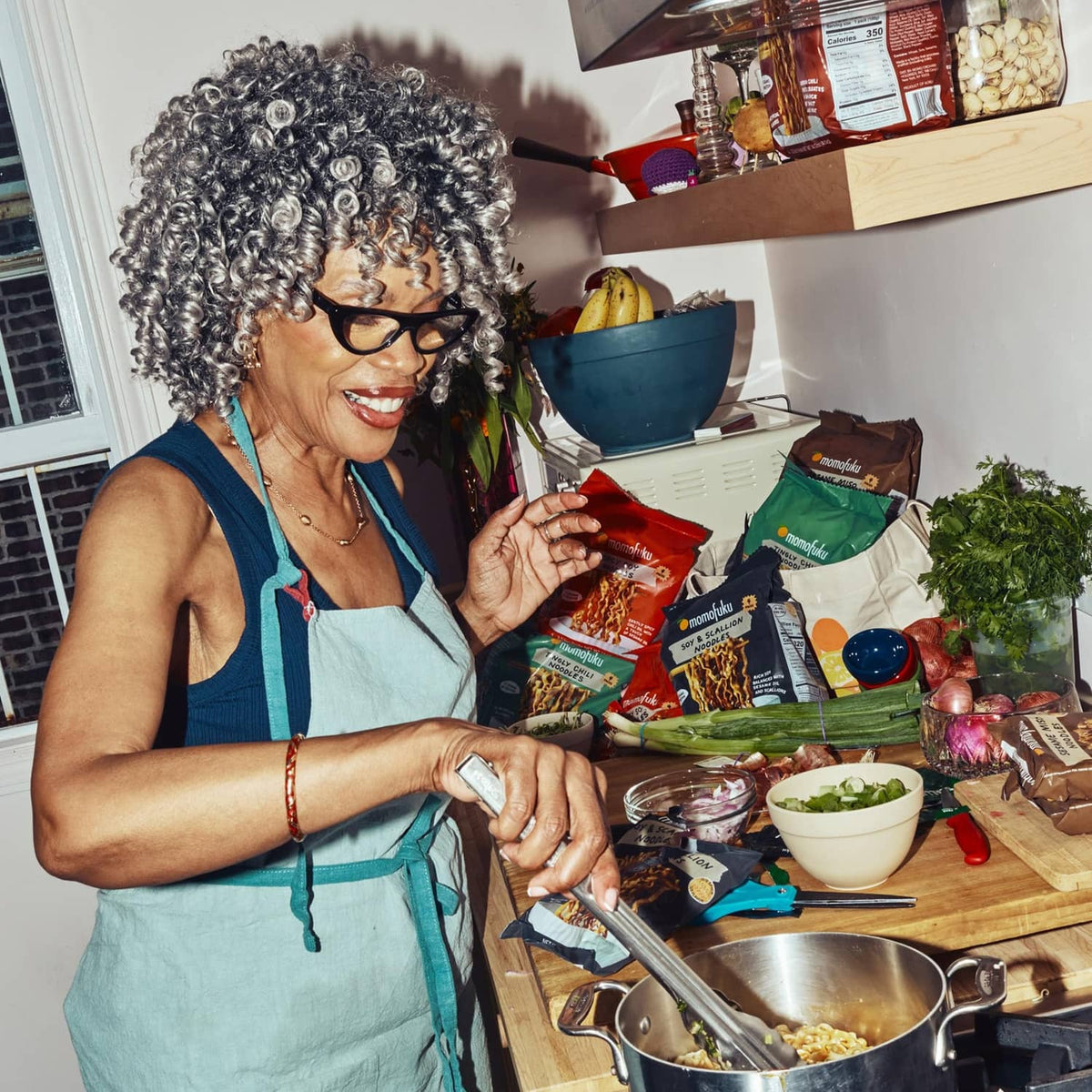 Momofuku ramen noodle packs on table beside woman cooking noodles in a pot