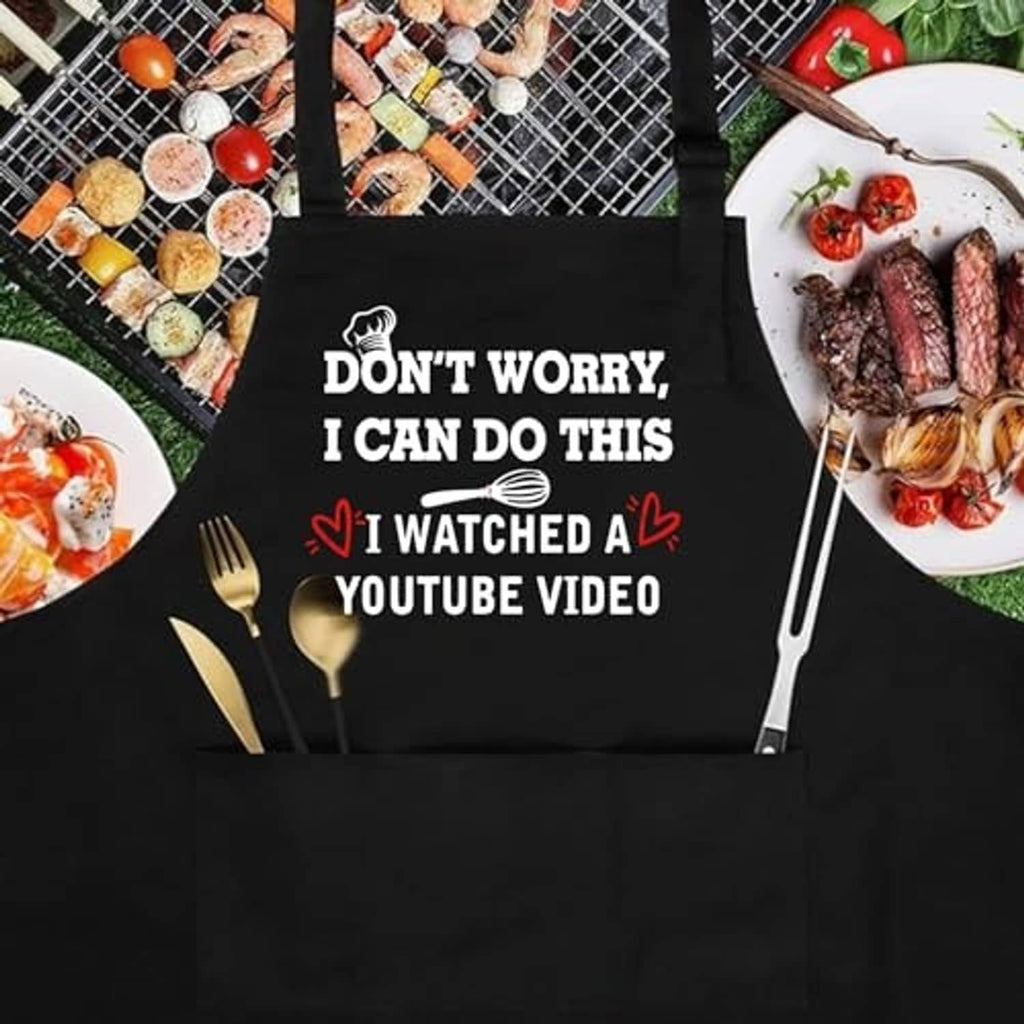 Black apron with "Don’t Worry, I Can Do This" slogan laid flat with cooking utensils nearby.