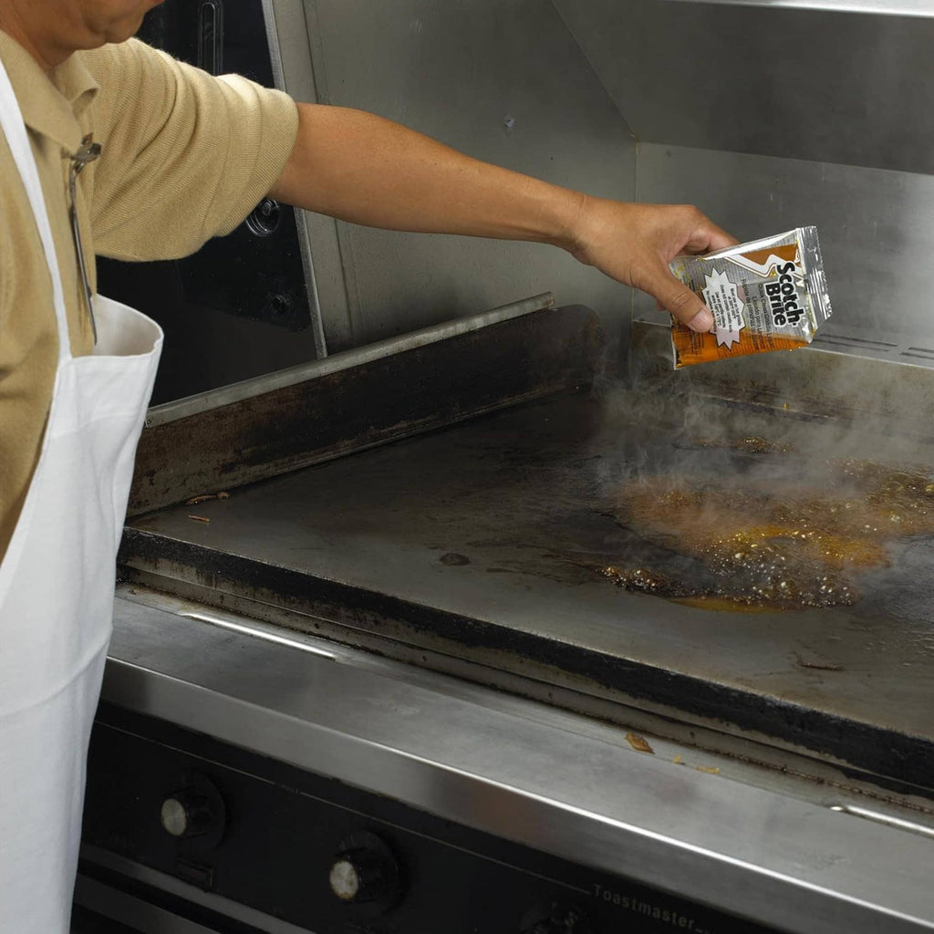 Scotch-Brite griddle cleaning liquid packet held above a metal flat-top griddle