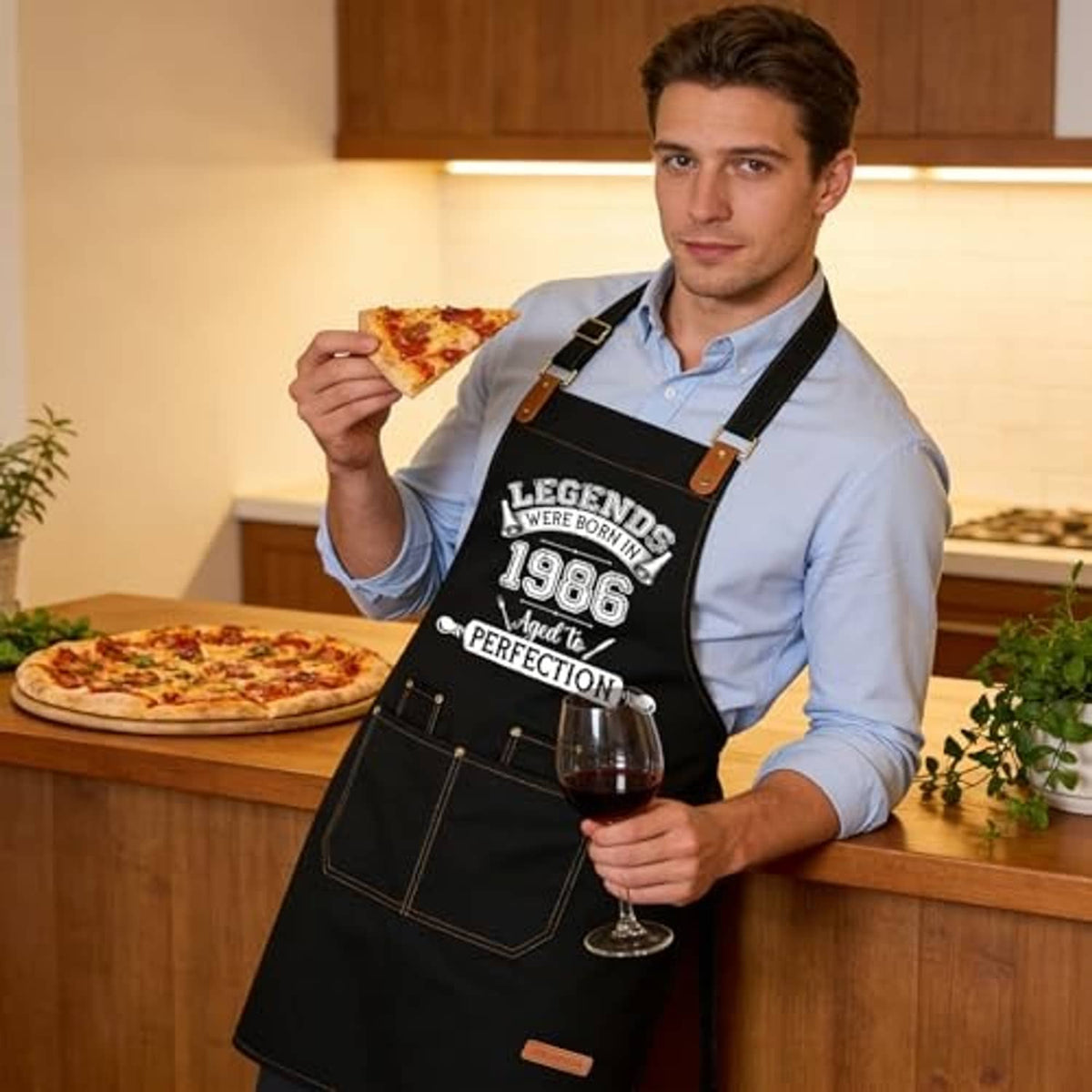 Black cooking apron with printed 1986 design, worn by a man holding a pizza slice.