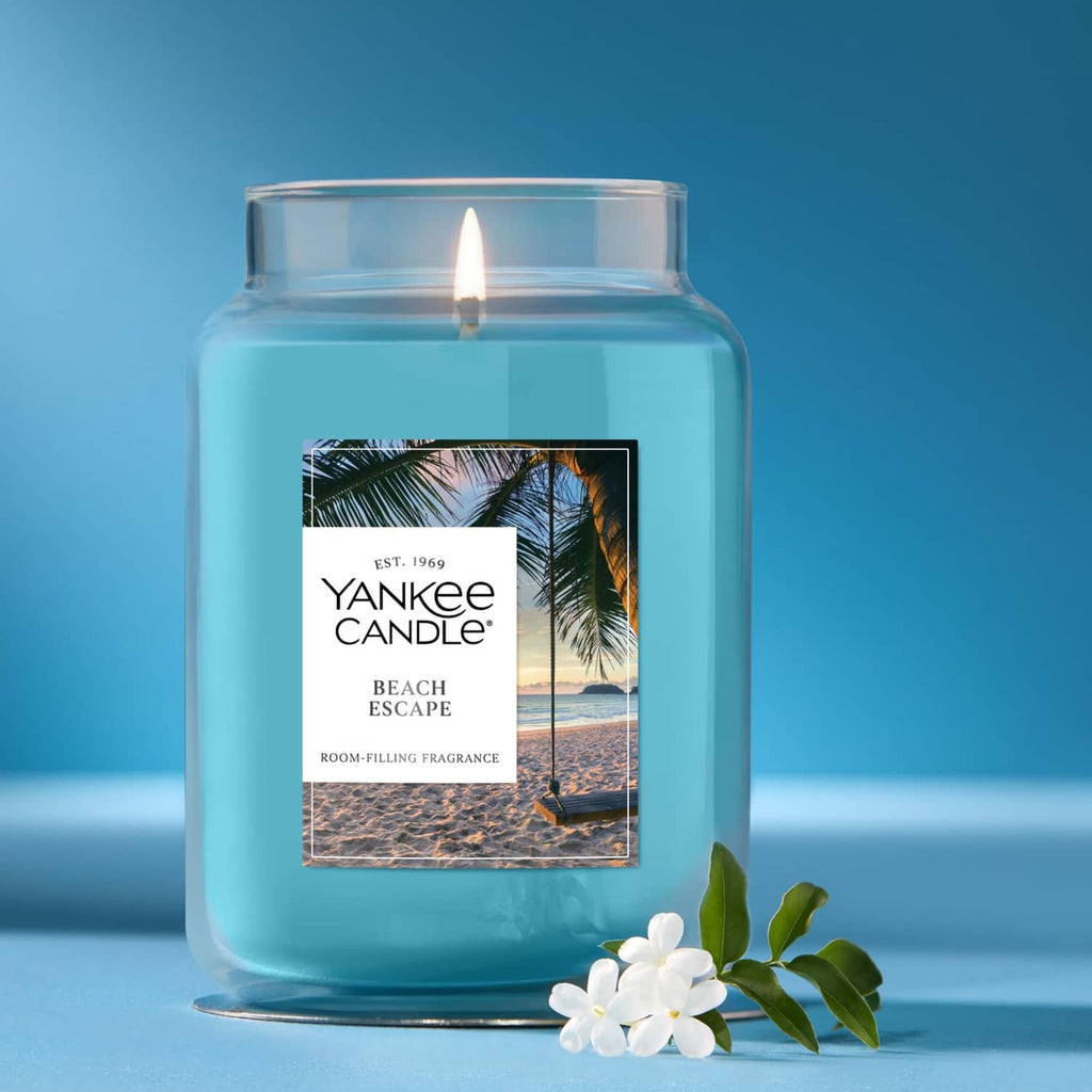 Blue glass jar candle with lit wick, white flowers, and Beach Escape label