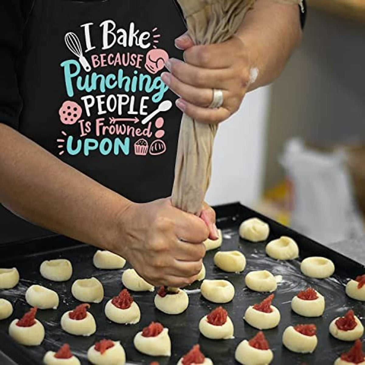 Black baking apron with 'I Bake' slogan worn while arranging dough on a tray
