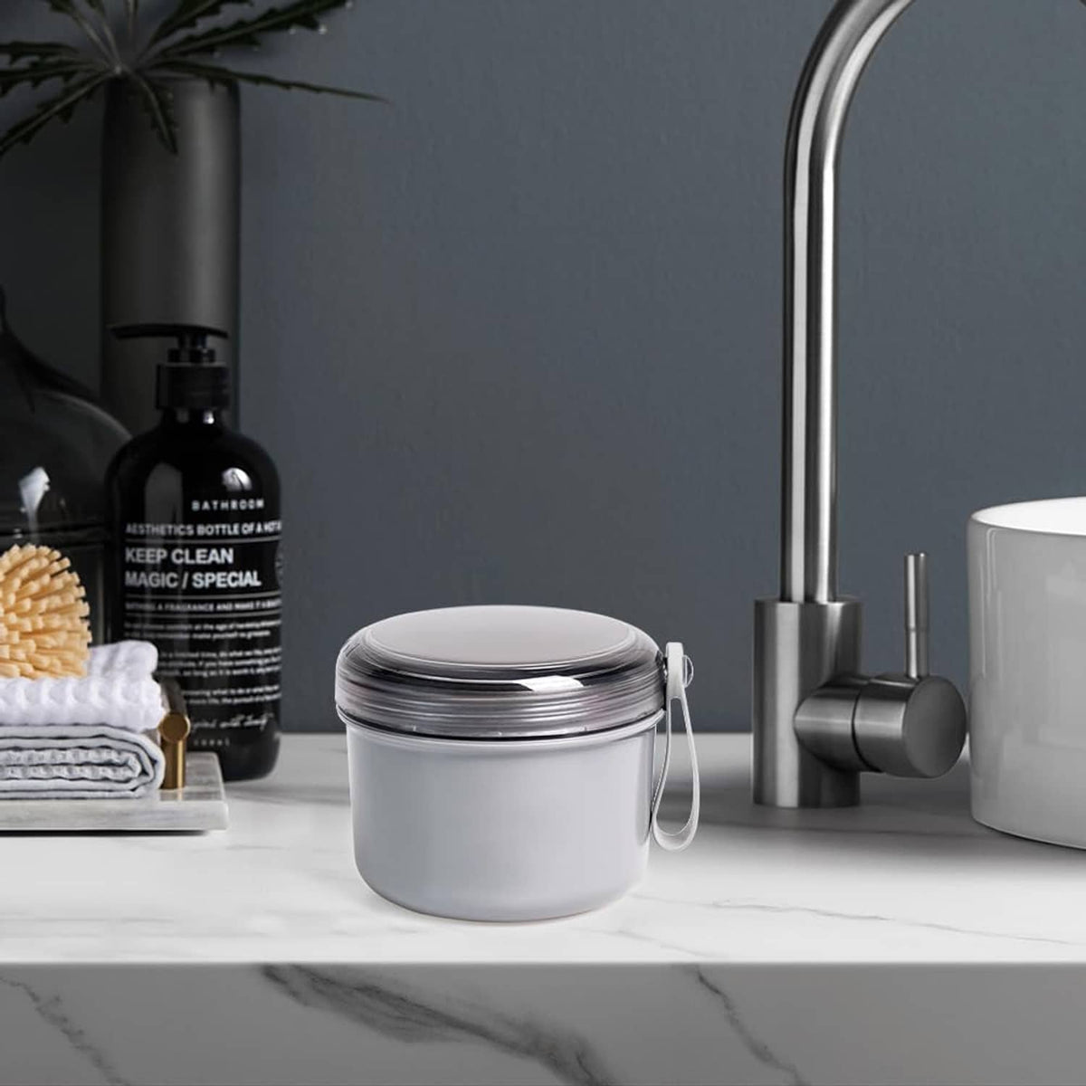Gray denture soaking cup with closed lid and hanging handle on countertop.