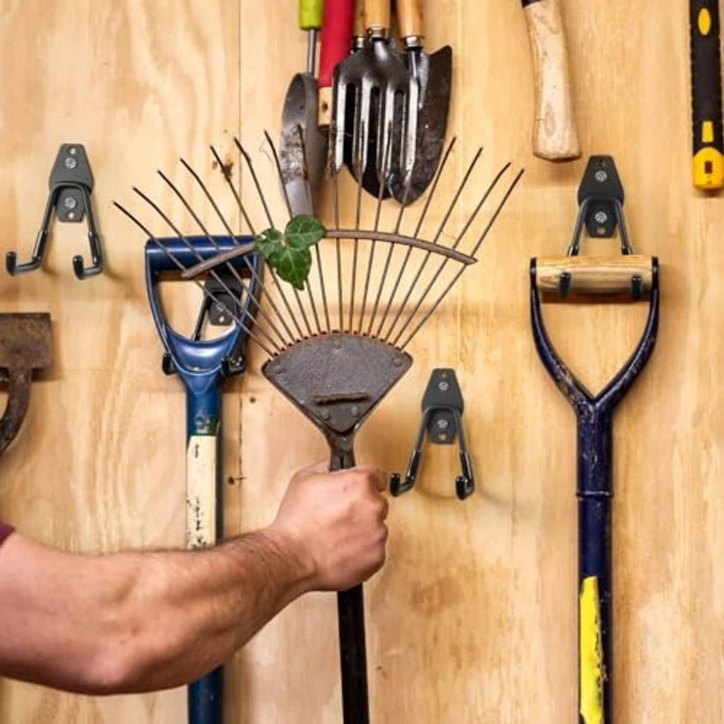 Black garage hooks mounted on a wooden board holding tools like shovels.