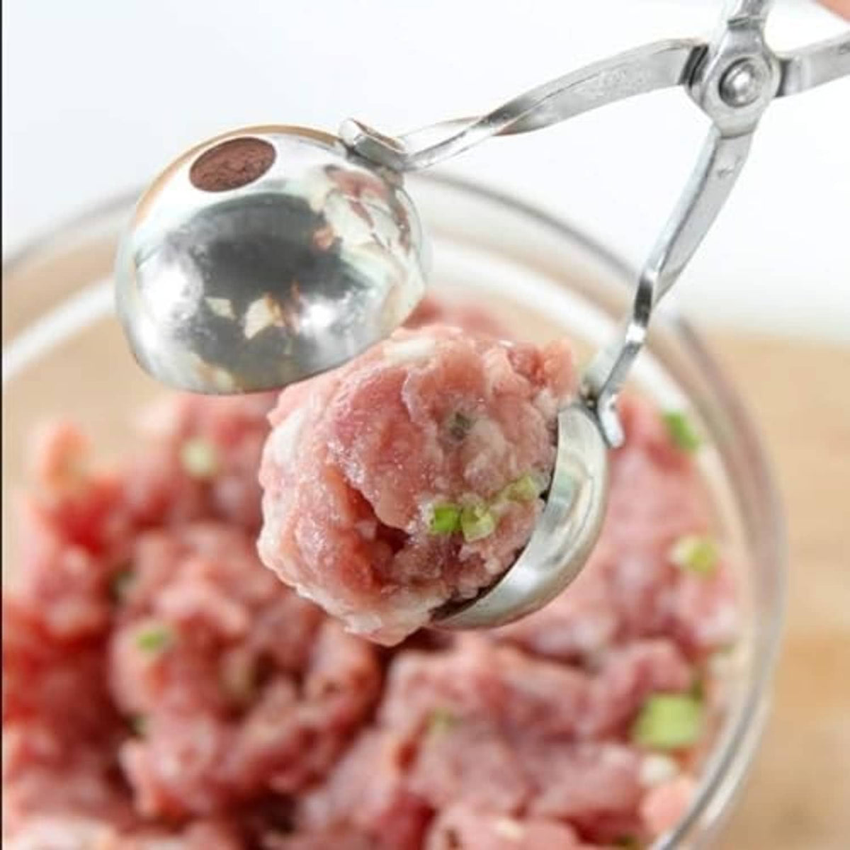 Stainless meatball maker scoop shaping raw meat into a ball over a mixing bowl.