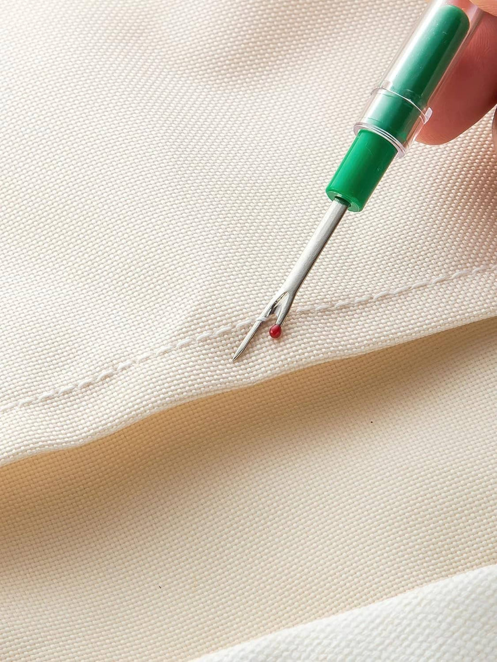 Green seam ripper tool lifting a stitch on beige fabric
