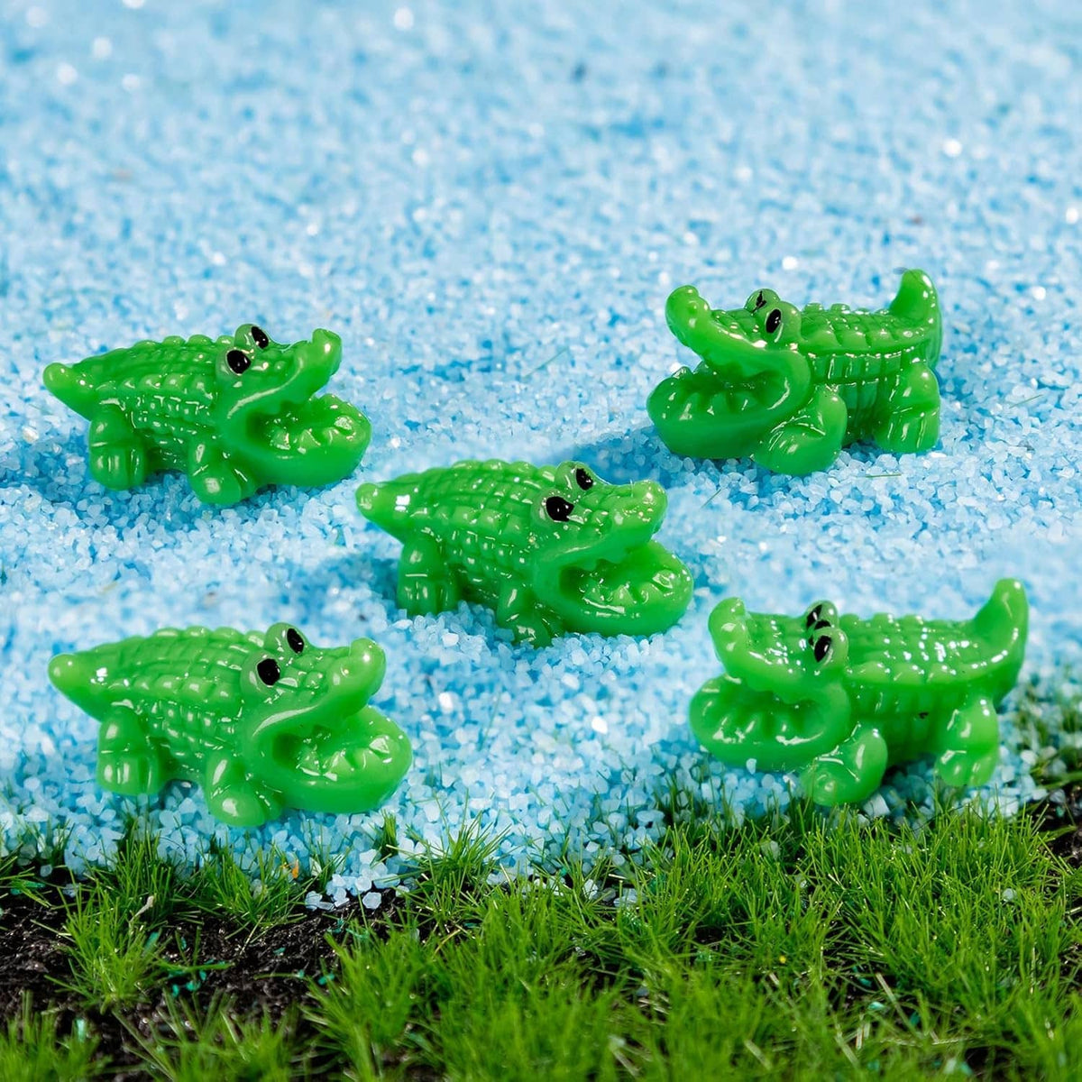 Assorted green mini crocodile resin figurines on blue gravel with grass nearby