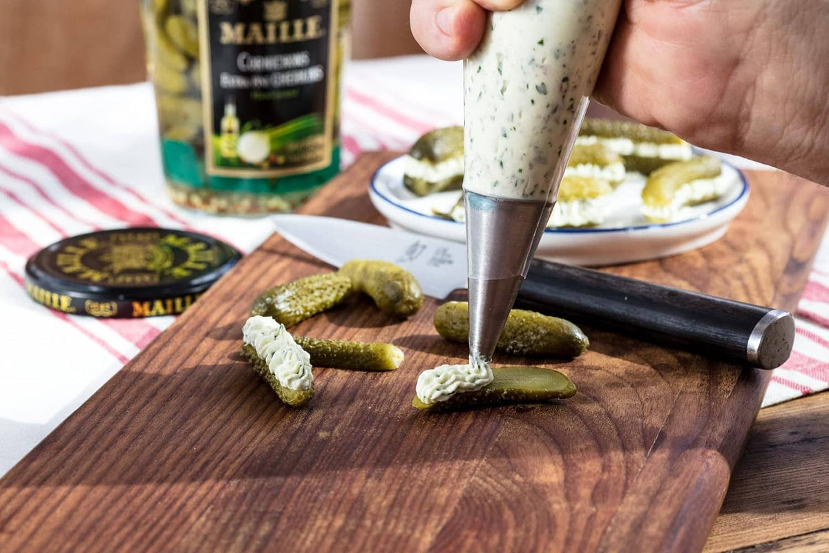 Hand piping creamy spread onto bread topped with green cornichons on board