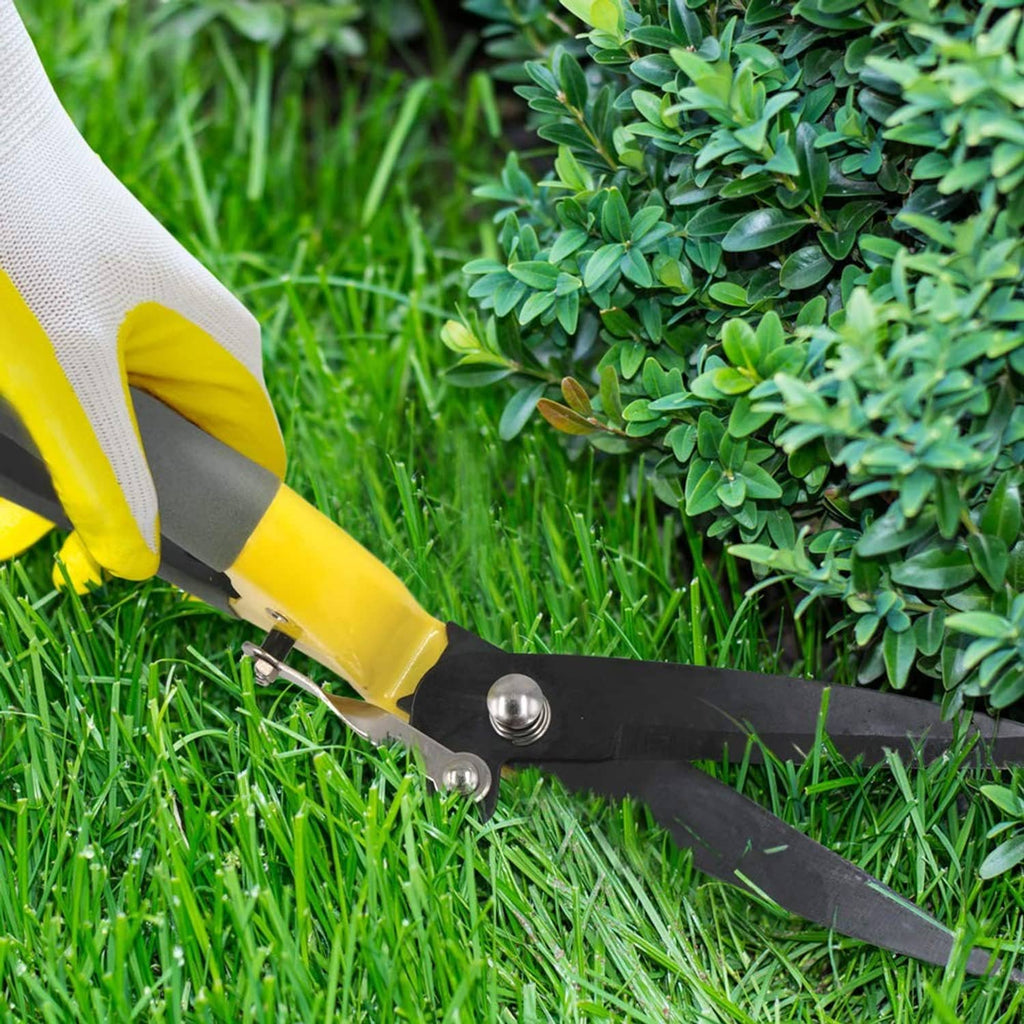 Handheld grass shears cutting along green shrub leaves, yellow handle with gray blades.