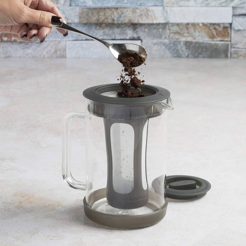 Glass cold brew coffee maker with black lid and handle, grounds being poured in