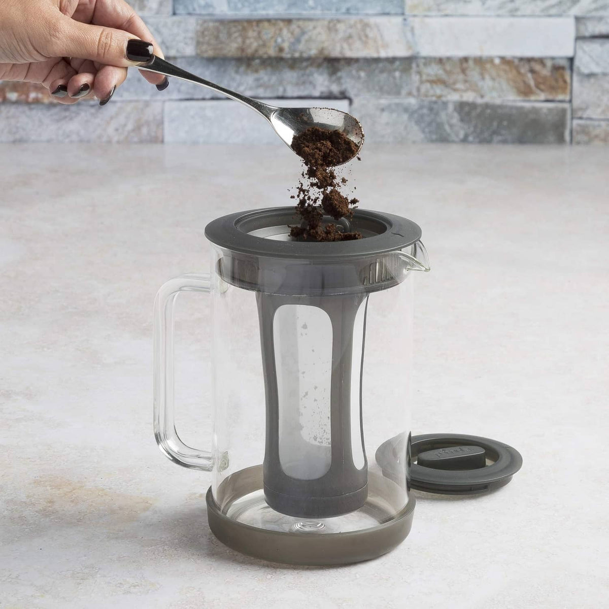 Glass cold brew coffee maker with black lid and handle, grounds being poured in