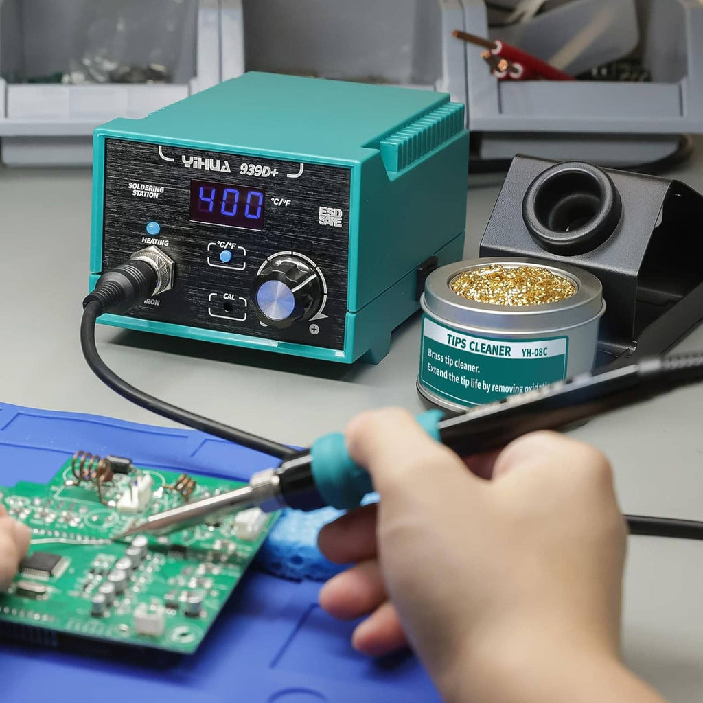 Digital soldering station with teal iron in use, hand soldering a circuit board, with tip-cleaner base