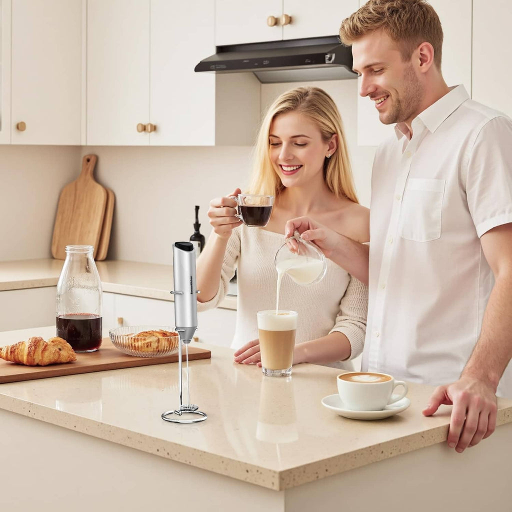 Silver milk frother on stand on countertop with glasses of frothed milk