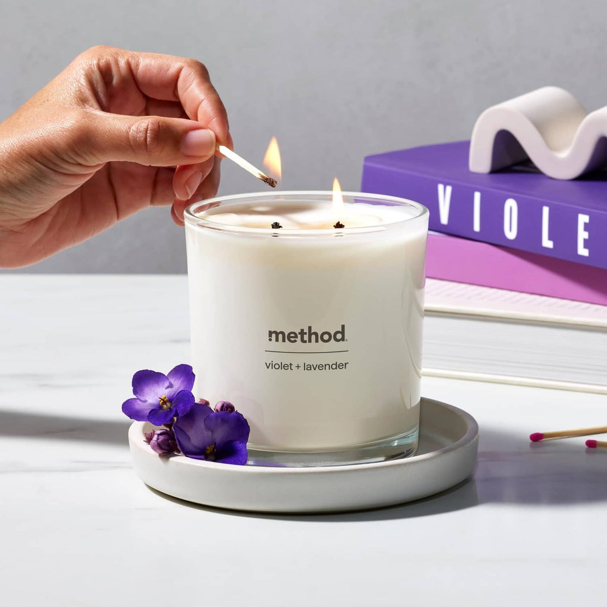 Lit violet + lavender soy candle in glass jar with two wicks, hand lighting