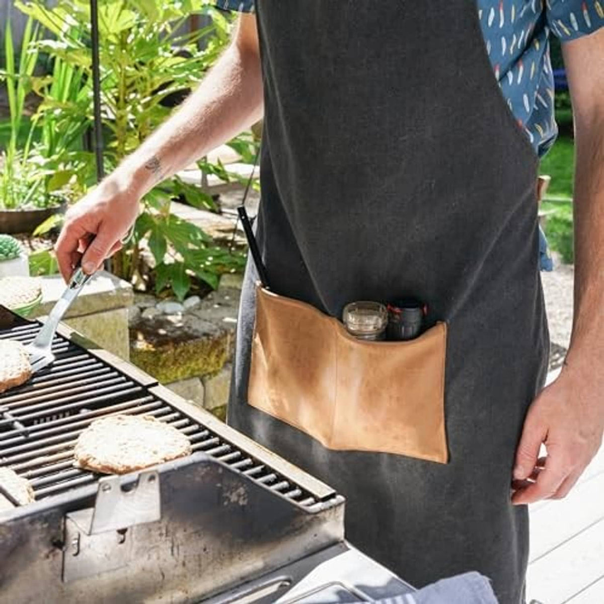 Dark charcoal apron with tan pocket holding small grilling tools and bottles