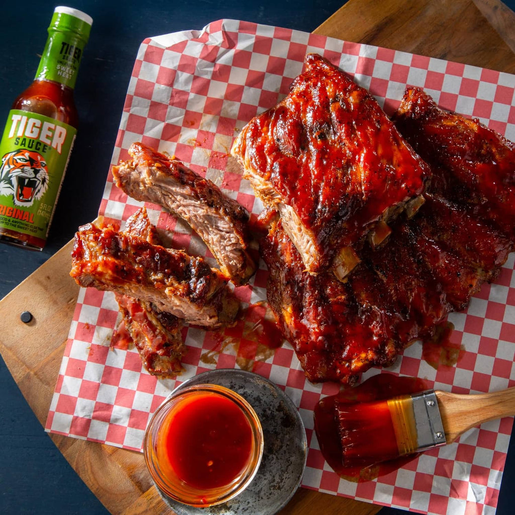 BBQ ribs with dark red glaze, green-capped hot sauce bottle and glass bowl