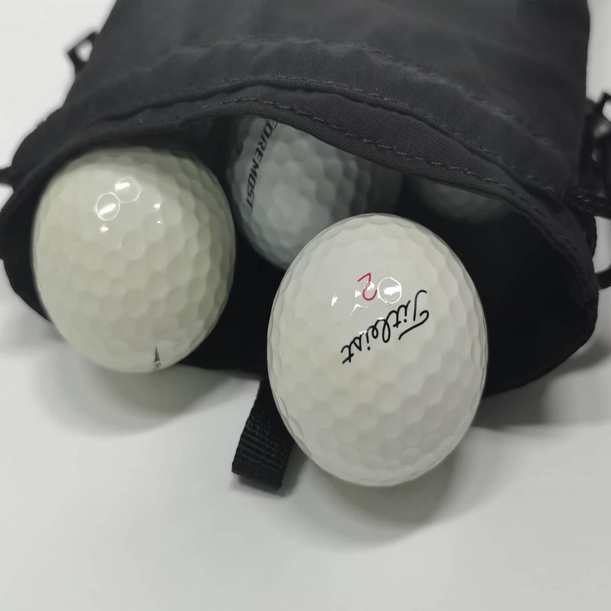 Black drawstring pouch holding three golf balls, with white print on the balls.