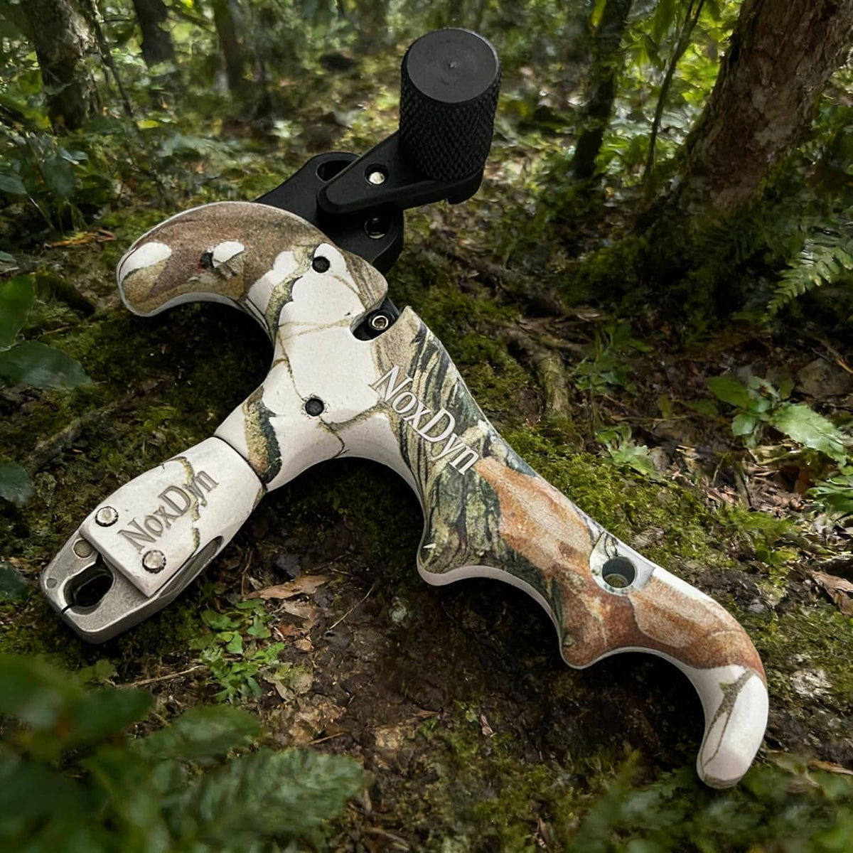 Archery bow thumb release with camo handle and black trigger, lying on forest floor