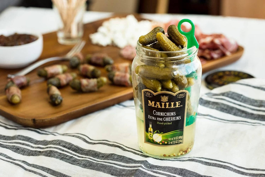 Jar of green cornichons with pickles on a wooden board at a meal table