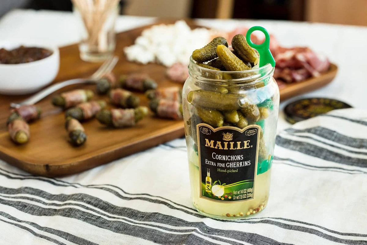 Jar of green cornichons with pickles on a wooden board at a meal table
