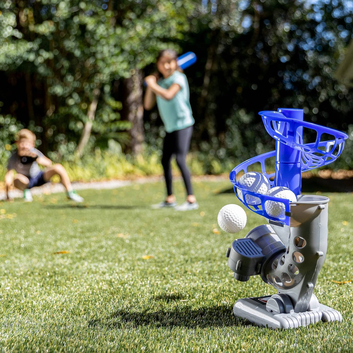 Blue and gray electronic pitching machine standing on grass with a stack of white balls