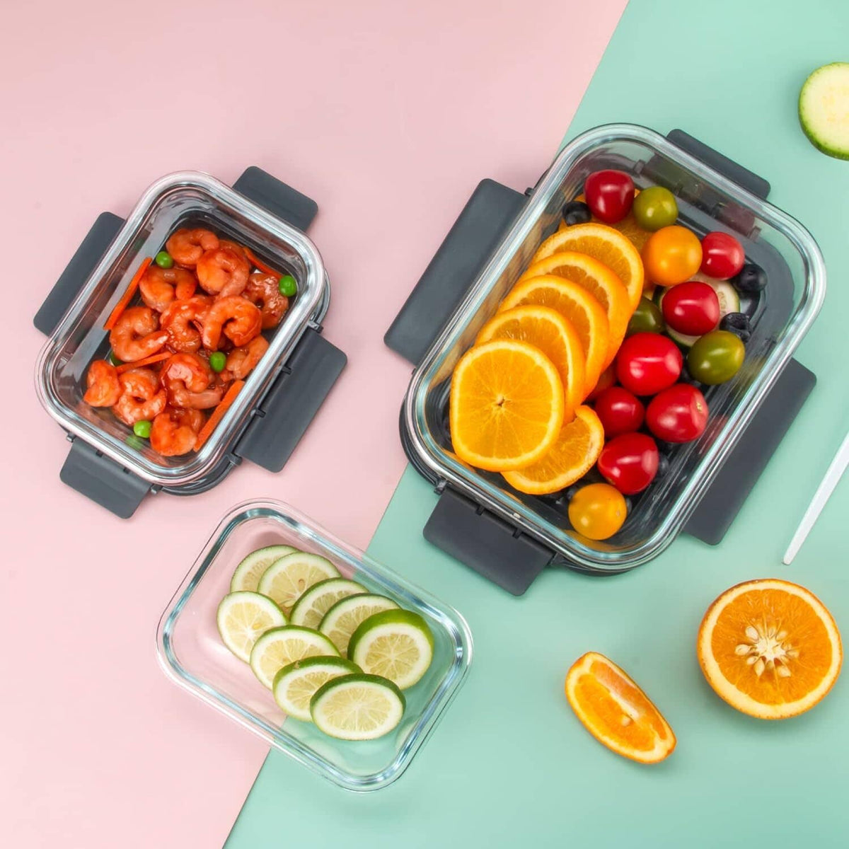 Glass food storage containers with dark lids filled with shrimp, fruit, and citrus