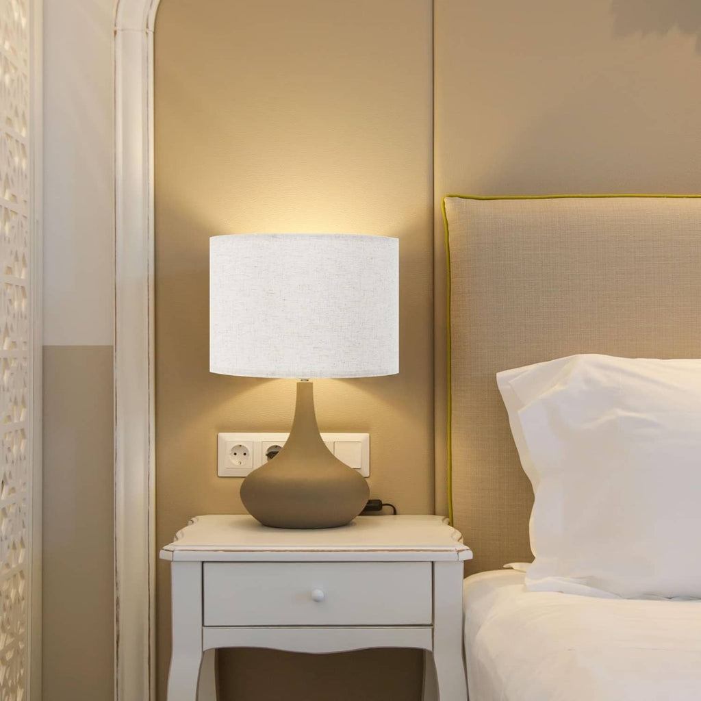 Beige linen drum lamp shade on table lamp base beside a pillow and nightstand