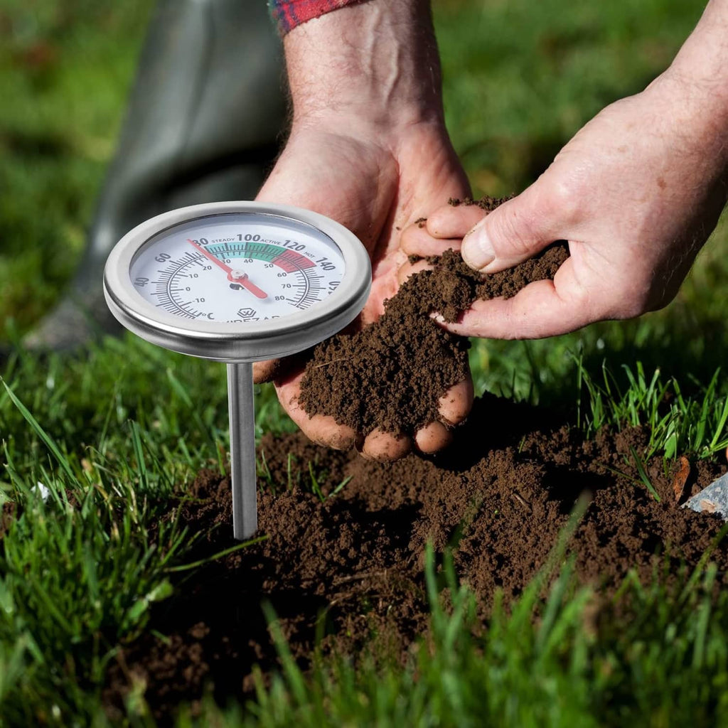 Soil thermometer with round dial on stainless probe, held by hands in dark soil