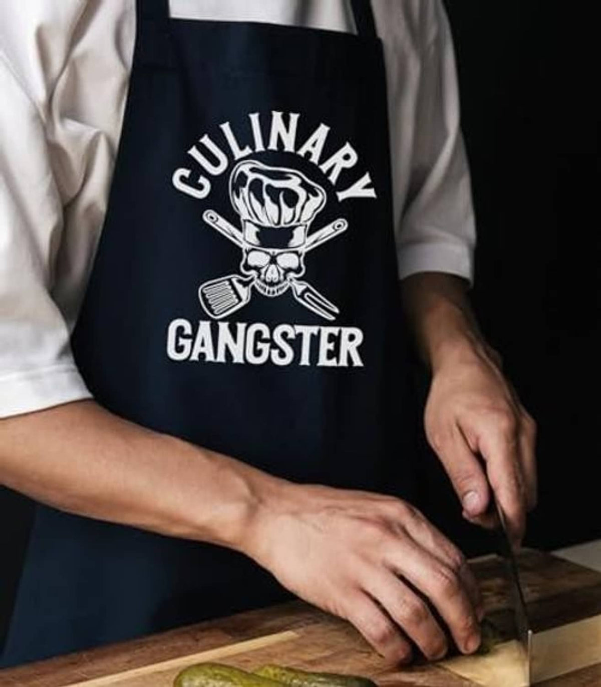 Black apron with skull and utensils graphic, worn while cutting on a wooden board.