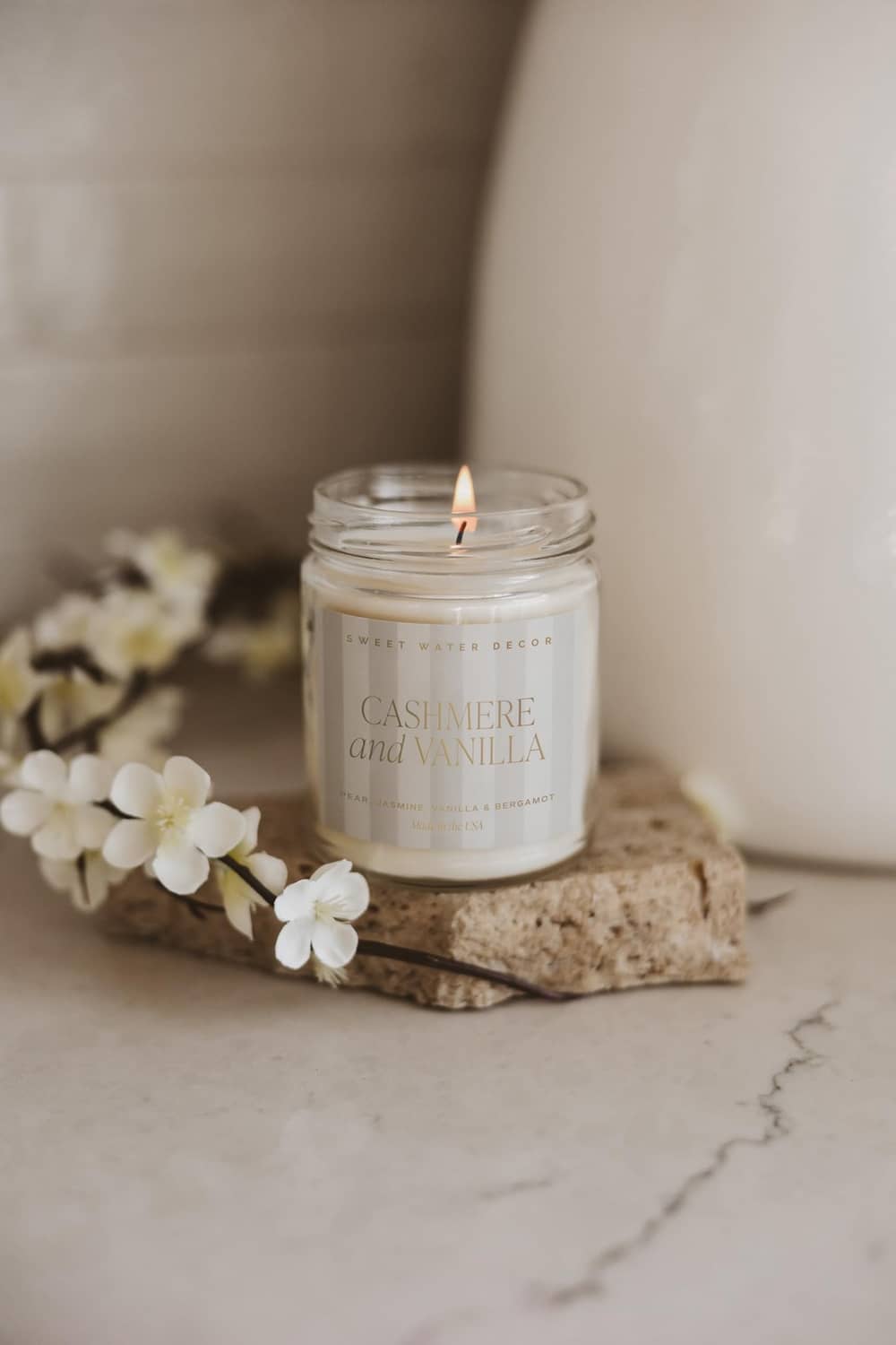 Small lit cashmere and vanilla candle jar with white flowers on a stone surface