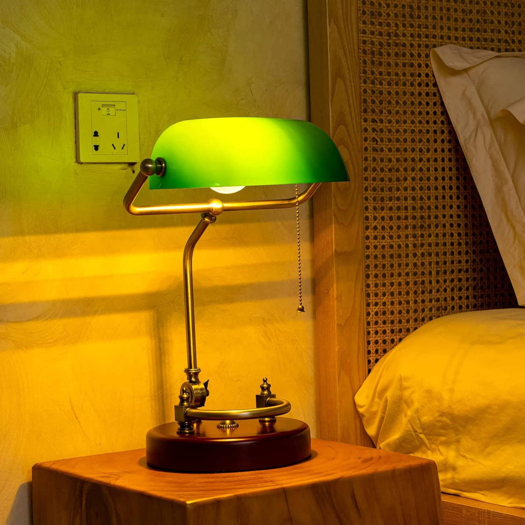 Green glass bankers desk lamp with brass frame and round red base on wooden table
