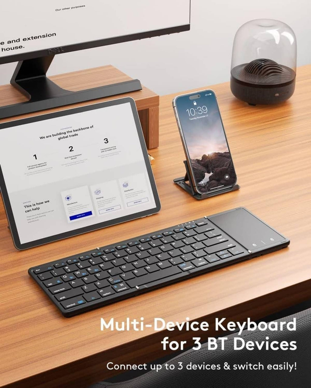 Black foldable Bluetooth keyboard with touchpad on desk, with tablet and phone nearby