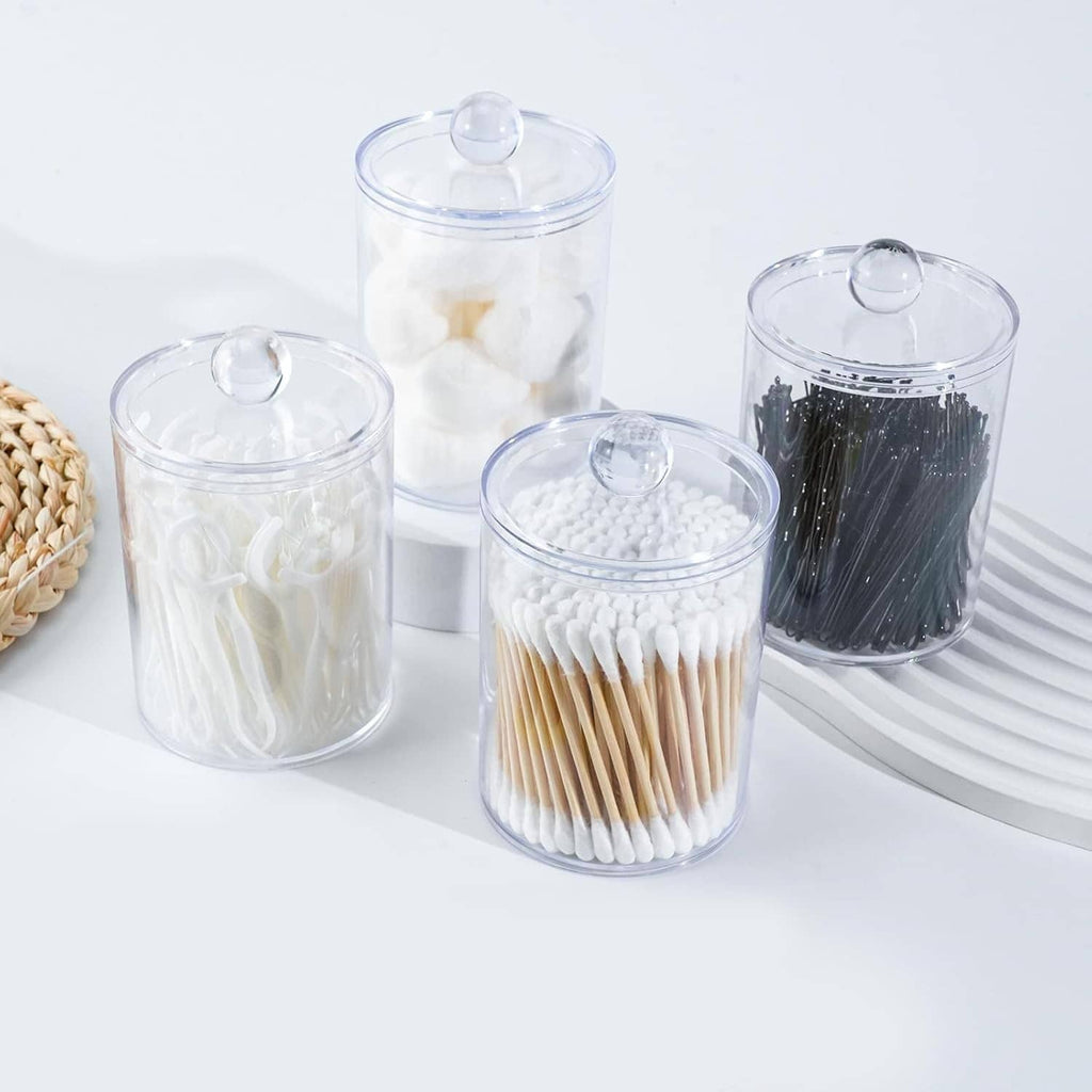 Four clear apothecary canisters with lids holding cotton swabs, cotton pads, and black accessories